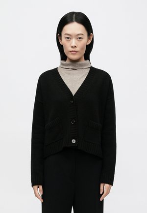 Cardigan noir en tricot avec col en V, bords côtelés, deux poches avant et fermeture à boutons. Porté par-dessus un pull à col roulé beige.