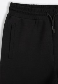 Czarne dresy o gładkiej teksturze, elastycznym pasie i regulowanym sznurku. Posiadają boczną kieszeń o eleganckim designie.
