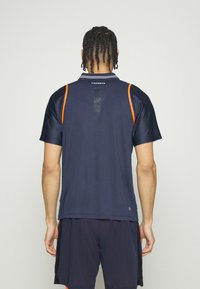 Marinblå idrottströja med korta ärmar, mesh-detaljer, orange accent och en logotyp bakpå. Lätt och ventilerande tyg.