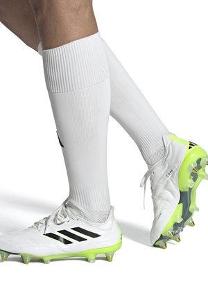 White soccer cleats with black stripes, neon yellow accents, and a textured surface, paired with ribbed white socks, showing leg.