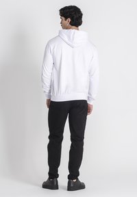 Biela mikina s klokaním vreckom a šnúrkou, skombinovaná s čiernymi slim-fit nohavicami a čiernymi topánkami, pohľad zozadu.