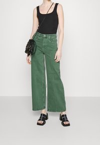 Jeans anchos verdes de denim con bolsillos delanteros y costuras visibles. Combinados con una blusa negra sin mangas y un bolso negro.