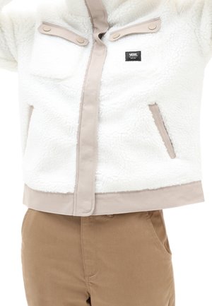 Witte fleecejas met beige accenten, voorzakken met drukknopen en een ritssluiting. Bruine broek met steekzakken aan de zijkant, gemaakt van stevige stof.