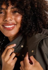 Chaqueta verde oscuro con un gran cuello que presenta broches a presión dorados, costuras visibles y una textura suave, llevada por una persona sonriente.