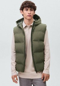 Olive zeleno podložena brezrokavnik s kapuco, z zadrgo in teksturirano tkanino. Nosijo ga čez svetlo bež srajco s ovratnikom.