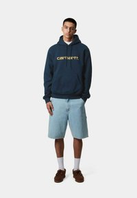 Sudadera azul marino con texto amarillo, material de algodón, bolsillo frontal, acompañada de pantalones cortos de mezclilla claros y zapatos marrones. Calcetines blancos visibles.