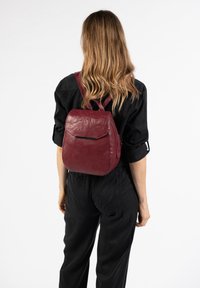 Roter Leder-Rucksack mit runder Form, kleiner Fronttasche und verstellbaren Riemen. Glatte Textur und minimalistisches Design.