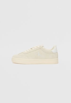Beige suede sneakers with a low-top design, lace closure, and light-colored accents. Embossed logo and textured midsole detail.