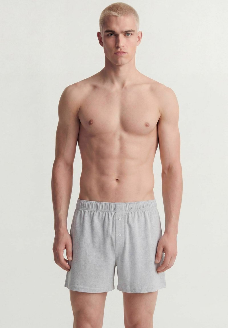 Jeune homme aux cheveux blonds courts portant un shorty en coton gris clair, debout devant un fond blanc uni.