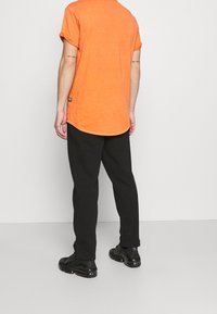 Oranžové bavlněné tričko s ohrnutými rukávy, zakřiveným lemem a malým štítkem značky na levé straně, spojené s černými volnými kalhotami a černými teniskami.