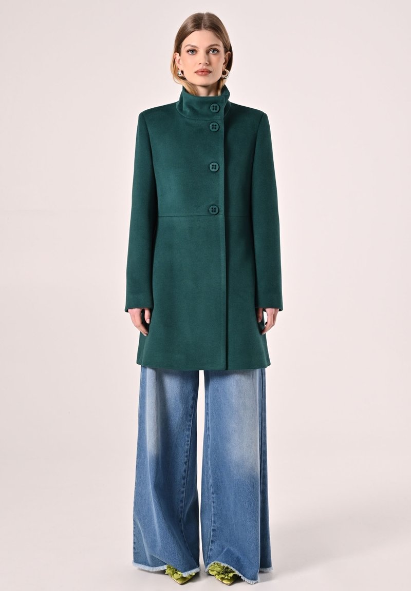 Donkergroene, knielange jas met een hoge kraag en zes knopen, gecombineerd met brede lichtblauwe spijkerbroeken en groene accent schoenen.