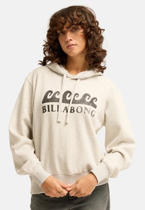 Donna con capelli ricci che indossa una felpa con cappuccio beige chiaro con grafiche di onde e testo "BILLABONG", in piedi con le mani vicino alla vita su sfondo uniforme.