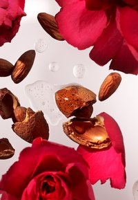 Mandeln und Rosenblätter, die in Wassertropfen schweben; braune und pinkfarbene Farben, glatte und strukturierte Oberflächen, mit sichtbaren gespaltenen Mandelschalen.