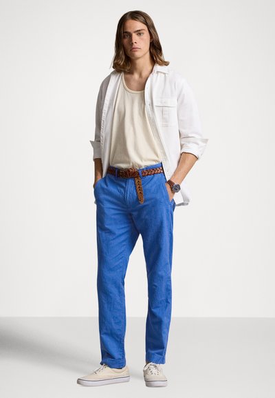 Μπλε λινά παντελόνια με πλεγτή καφέ δερμάτινη ζώνη, λευκό πουκάμισο με κουμπιά και μπεζ αθλητικά παπούτσια. Απαλή υφή και άνετη εφαρμογή.