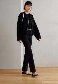 Crna oversized flis jakna sa zatvaračem, crne uske traperice i siva sandala. Drveni pod i svijetlosiva zidna pozadina.