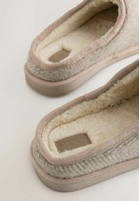 Beige čības ar vilnas ārpusi un mīkstu mākslīgo kažokādu iekšpusē. Piedāvā noapaļotu pirkstu un teksturētu rakstu, ar plakanu gumijas zoli.