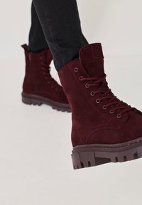 Botine din suede burgundy, cu limbă căptușită, vârf rotunjit, închidere cu șireturi și talpă de cauciuc groasă, cu striații pronunțate.