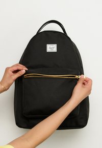 Zaino in tessuto nero con parte superiore arrotondata, tasca frontale con zip dorata e patch logo bianco. Manici per un facile trasporto.