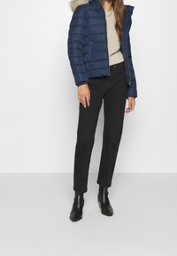 Kvinna klädd i en marinblå puffjacka med pälsHuva, beige stickad tröja, svarta byxor och svarta ankelstövlar som står framför en neutral bakgrund.