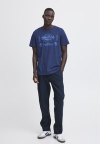 Tumši zila kokvilnas T-krekls ar grafisku apdruku un gaismas zila tekstu, savienots ar tumšām biksēm un baltām kedām ar melnām svītrām.