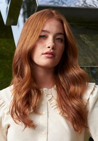 Jeune femme aux longs cheveux roux ondulés et taches de rousseur, portant un chemisier crème avec un col à volants, posant à l'intérieur devant un fond métallique réfléchissant.