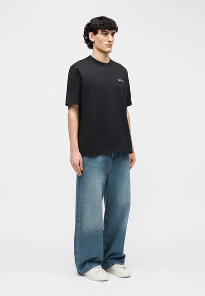 Νεαρός άνδρας που στέκεται φορώντας μαύρο t-shirt, άνετα μπλε τζιν και λευκά αθλητικά παπούτσια, μπροστά σε ένα απλό φωτεινό φόντο.