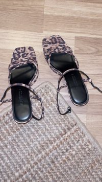 Et par leopardmønstrede sandaler med åben tå og ankelremme, placeret på trægulv ved siden af hjørnet af et tekstureret beige vævet tæppe.