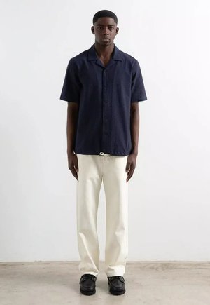Jeune homme debout, portant une chemise à manches courtes bleu marine, un pantalon blanc à cordon et des chaussures noires sur un fond clair uni.