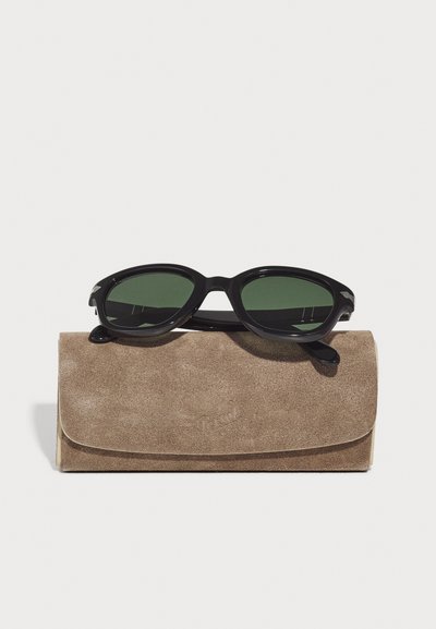Occhiali da sole neri con lenti verdi poggiati su una custodia in suede color tan. La custodia ha un flap arrotondato e un logo in rilievo sottile.