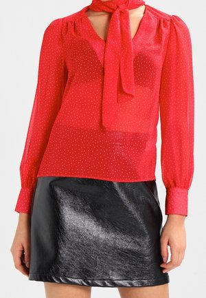Blouse rouge à manches longues avec petits pois blancs et un nœud au cou, associée à une mini-jupe en cuir noir brillante.