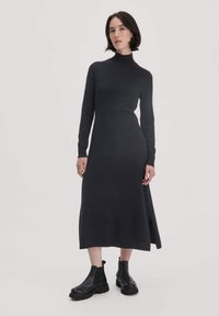 Frau steht in einem langen schwarzen Rollkragenkleid mit Seitenschlitz und schwarzen klobigen Stiefeletten vor einem schlichten Hintergrund.