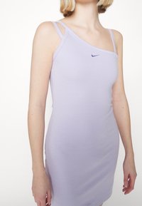 Slim-fit ljuslila klänning med dubbel axelremmar och en liten mörk Nike-logotyp på bröstet, bärs av en person med kort blont hår.