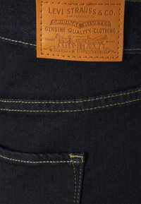 Jeans de mezclilla oscura con un parche de cuero marrón con marca en relieve, costuras amarillas y un detalle de remache.