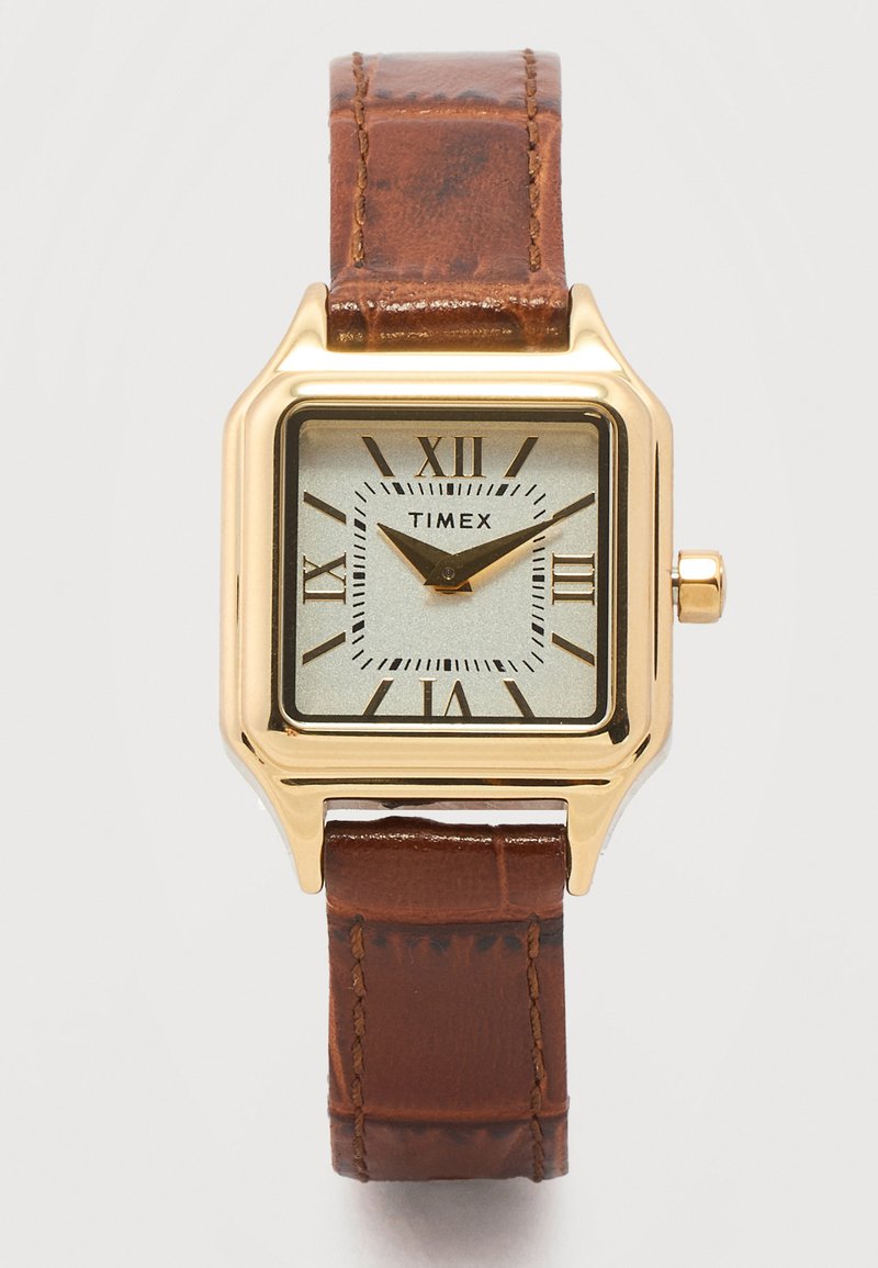 Τετράγωνο ρολόι Timex σε χρυσή απόχρωση με ασημί καντράν, μαύρους λατινικούς αριθμούς, μαύρα δείκτες και καφέ δερμάτινο λουράκι με ραφές.