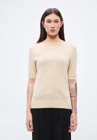 Pulover bej cu mâneci scurte, confecționat din țesătură moale tricotată, cu decolteu rotund și tiv cu refile, purtat cu pantaloni negri.