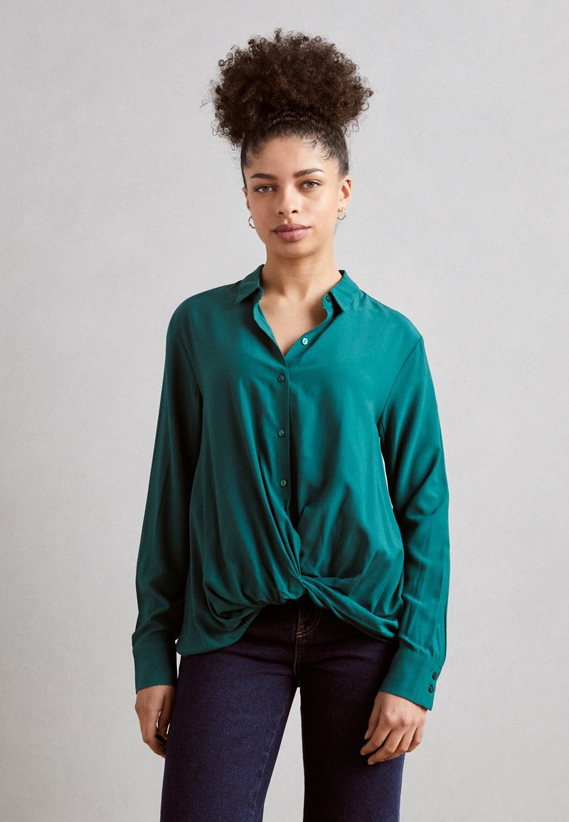 Camicia verde a maniche lunghe con bottoni, dotata di un dettaglio a nodo torsionato nella parte frontale, realizzata in tessuto liscio e con colletto e polsini.