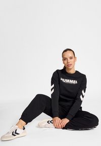 Czarna bluza z kapturem z logo "HUMMEL", białe akcenty w kształcie chevron na rękawach; zestawiona z czarnymi dresami i beżowymi sneakersami w czarne paski.