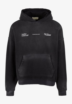 Haut à capuche noir en tissu doux avec une poche avant. Présente des logos en texte blanc et une capuche avec cordon de serrage. Les bords ont une finition brute.