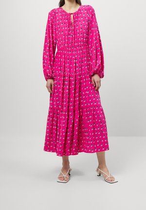 Vrouw draagt een felroze midi-jurk met lange mouwen, een klein bloemenpatroon en een keyhole-halslijn, gecombineerd met witte sandalen met hakken.