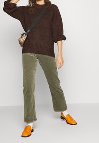 Personne portant un pull en maille marron, un pantalon en velours côtelé vert olive, des chaussettes blanches, des mocassins orange vif et un sac bandoulière imprimé léopard.