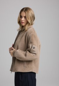 Beige Kunstfell-Bomberjacke mit Reißverschluss vorne, gerippten Bündchen und einer aufgesetzten Tasche an einem Ärmel. Strukturierte Oberfläche und lockere Passform.