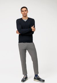 Marineblauwe gebreide trui met V-hals, grijze taps toelopende broek en zwarte sportschoenen met blauwe accenten, staande op een effen witte achtergrond.