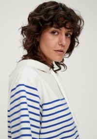 Weißer Hoodie mit blauen horizontalen Streifen, ausgestattet mit einer Kordelzugkapuze und einer weichen Textur. Das Kleidungsstück hat einen lockeren Schnitt und dezente Details.