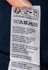 Étiquette d'entretien avec des instructions d'entretien en plusieurs langues. Matériau : 60 % coton, 40 % polyester. Texte blanc sur un fond en tissu bleu foncé.