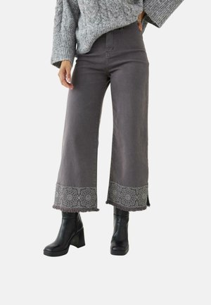 Mujer con pantalones grises anchos y cortados con puños bordados, botas negras de tacón y suéter gris de punto trenzado, posando frente a un fondo blanco.