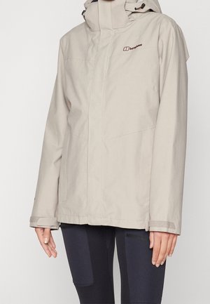 Beige waterdichte Berghaus jas met capuchon, voorzakken en klittenband polsbandjes, gedragen met zwarte wandellegging.