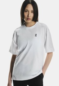 Ung kvinde med glat, mørkt hår iført en hvid oversized T-shirt med et lille sort logo på brystet, poserer med en hånd i lommen.