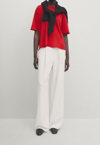 T-shirt rouge surdimensionné, pull noir drapé autour du cou, pantalon large blanc et chaussures noires. Design simple et couleurs unies.