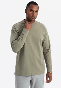 Mann, der sich am Hals fasst, trägt ein olivgrünes Langarmshirt und hellgraue Hose vor einem weißen Hintergrund.