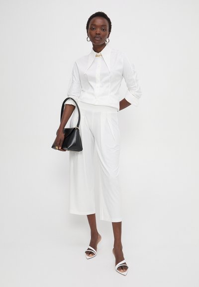 Weißes Hemd mit Kragen und weiße Culottes kombiniert mit weißen Absatzsandalen. Schwarze Handtasche mit goldenen Akzenten. Das Model steht vor einem hellen Hintergrund.